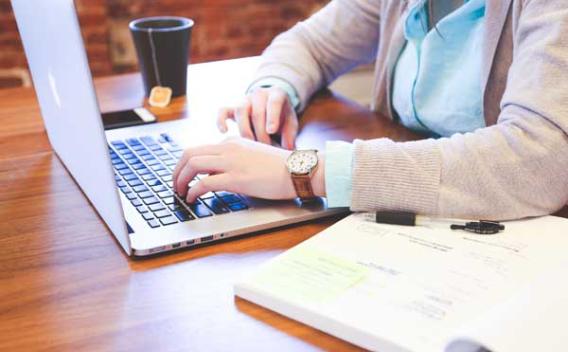 View of person's arms and hands on a laptop at a table with open book and teacup nearby